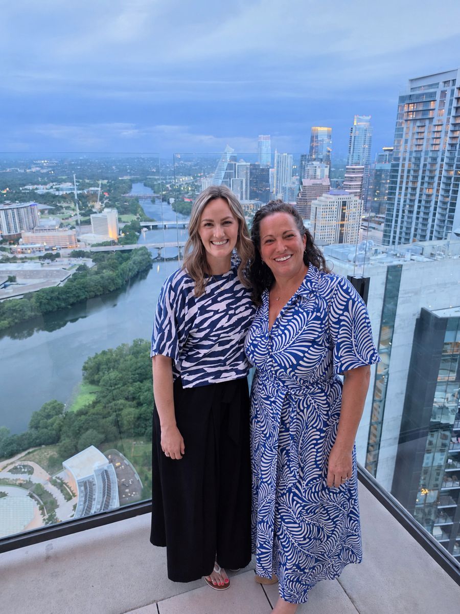 Shannon and Jessica standing close together and smiling on a balcony overlooking the Austin skyline at dusk
